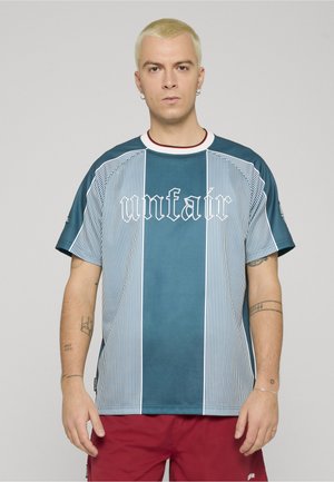 Mann med kort blondt hår iført en blåstripet skjorte med teksten «unfair» og røde shorts, stående foran en ensfarget grå bakgrunn.