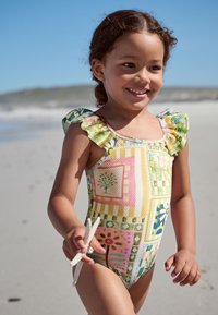 Badeanzug mit farbenfrohem Patchwork-Design, versehen mit Rüschenärmeln, texturiertem Stoff sowie Blumen- und geometrischen Mustern. Kind hält Muscheln.