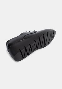Schwarze Sneakers mit einer glatten, strukturierten Oberfläche, ausgestattet mit einer dicken, schwarzen Gummisohle und einem Zick-Zack-Profil für besseren Grip.