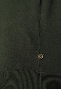 Tessuto a maglia ribbed di un verde scuro, con una superficie liscia. Presenta un piccolo accessorio a forma di logo circolare in tonalità oro vicino alla cucitura.