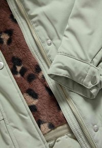 Giacca esterna verde con zip argentata, presenta un interno in finta pelliccia marrone con macchie di leopardo. Include bottoni a pressione sui polsini.