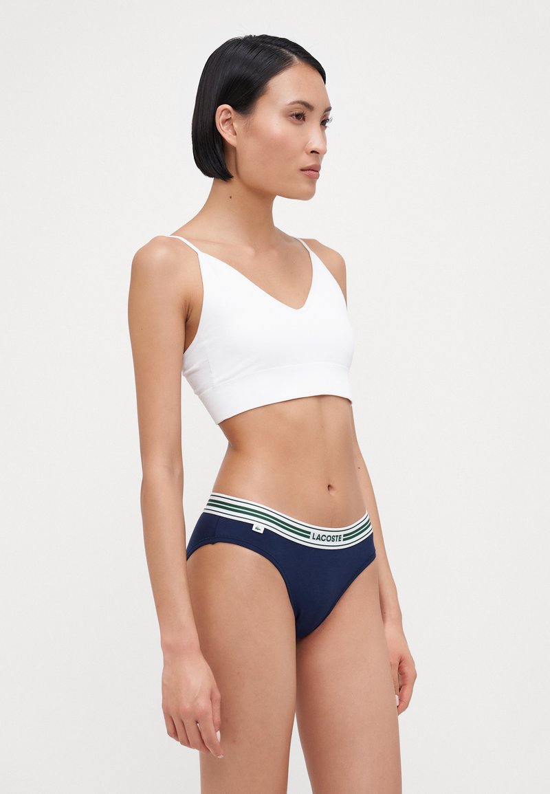 Bralette bianco a coste con scollatura a V abbinato a slip blu navy con un elastico verde e bianco con la scritta "LACOSTE."