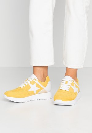 Stopy w żółtych sneakersach z białymi akcentami w kształcie gwiazdy i sznurowadłami, w zestawieniu z białymi, krótkimi spodniami, stojące na białej podłodze.