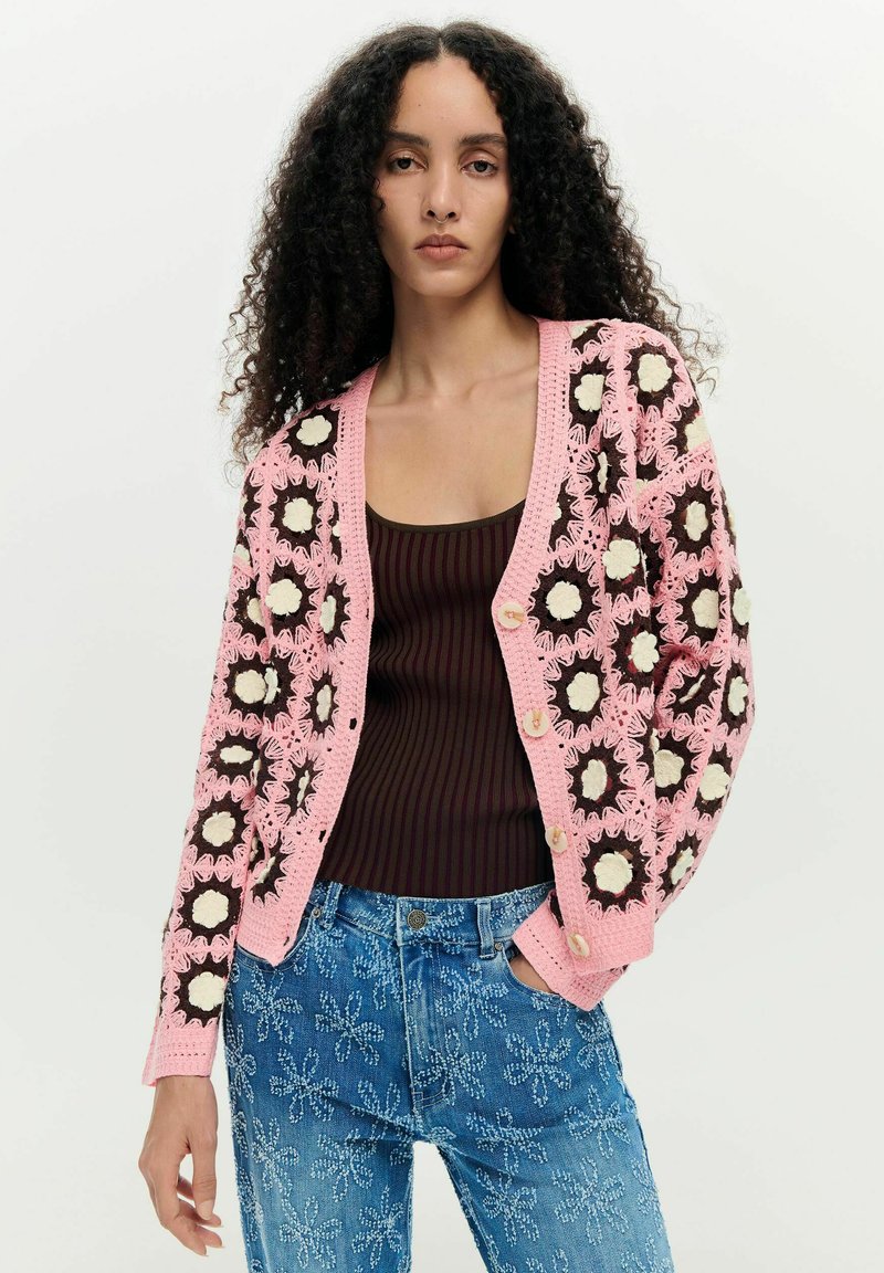 Femme aux longs cheveux bouclés portant un cardigan rose en crochet avec des motifs floraux, un débardeur rayé foncé et un jean bleu brodé de fleurs.
