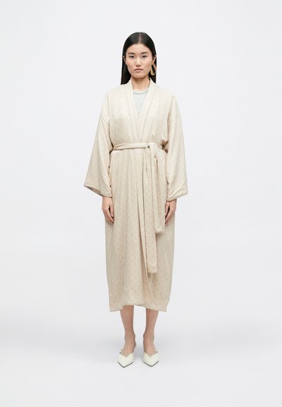 Μπεζ ριγέ ρόμπα με ζώνη στη μέση, μακριά μανίκια και ίσια γραμμή. Το μοντέλο φοράει λευκά μυτερά παπούτσια. Το φόντο είναι λευκό.