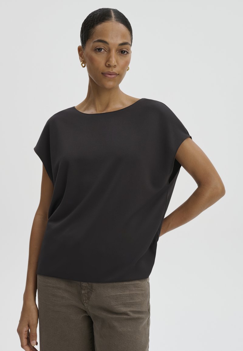 Woman with dark hair in a low bun wearing gold hoop earrings, a loose black short-sleeve top, and brown pants against a plain background.
