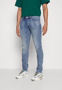 Obtažené světle modré džíny s roztržením na levém stehně, nošeny s zeleným tričkem a bílými teniskami s olivovými akcenty.