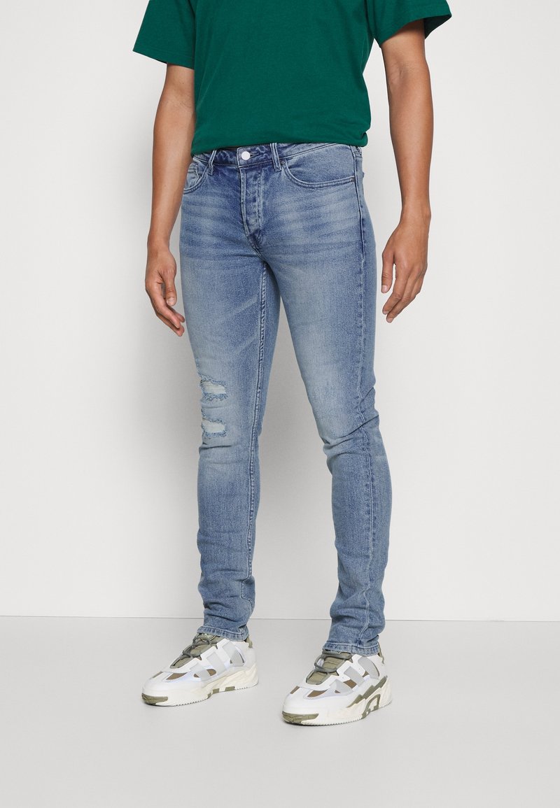 Obtažené světle modré džíny s roztržením na levém stehně, nošeny s zeleným tričkem a bílými teniskami s olivovými akcenty.