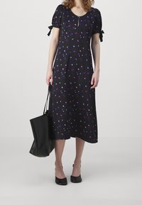 Kvinna i en svart midiklänning med lila blommönster, puffärmar med rosetter, svarta klackar och bär en stor svart tygväska.