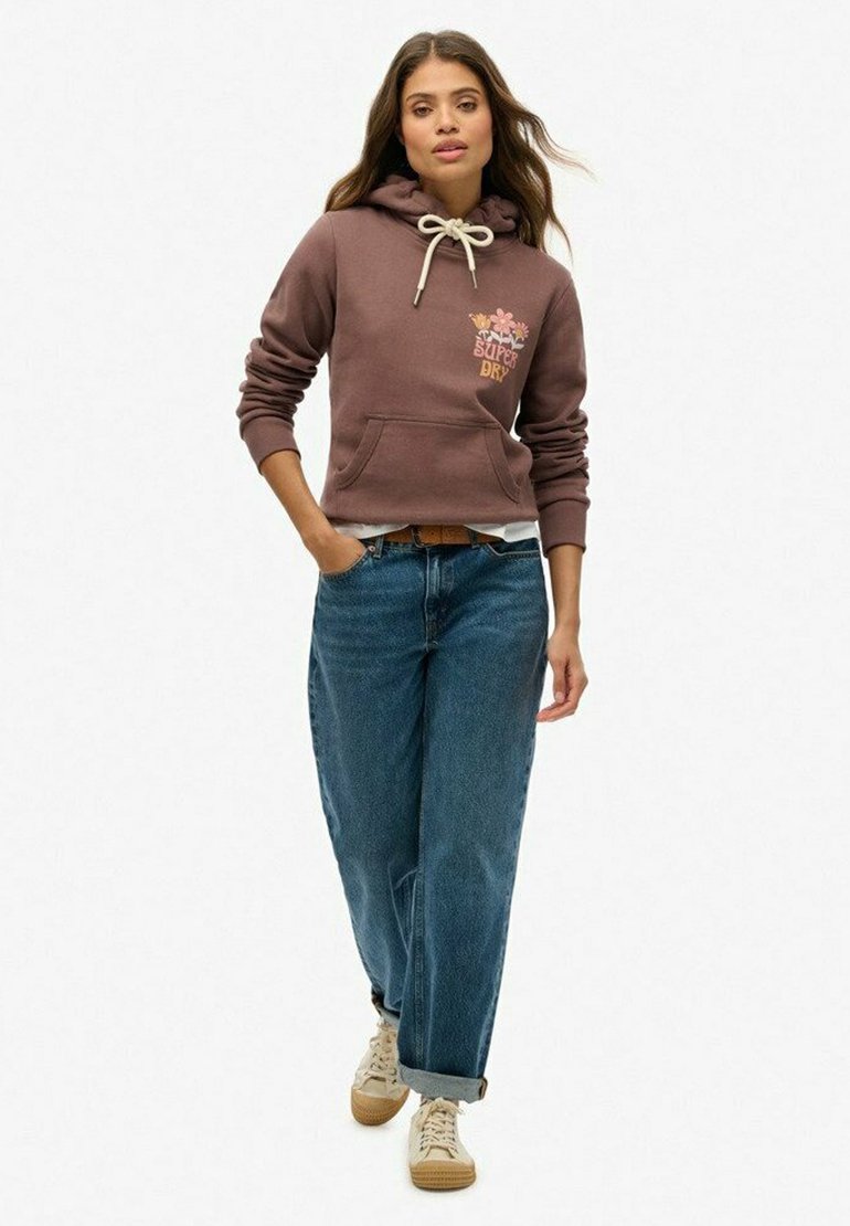 Hoodie marron avec cordon de serrage, doté d'un motif floral sur la poitrine. Associé à un jean bleu ample et des baskets beiges. Tissu texturé.