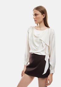 Hvid bluse med draperet front, lange ærmer og knapdetaljer, parret med en kort sort nederdel. Glat stof med en let glans.