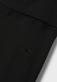 Czarne legginsy sportowe o gładkiej fakturze, z matowym czarnym detalem logo. Przeszycia są widoczne wzdłuż szwów.