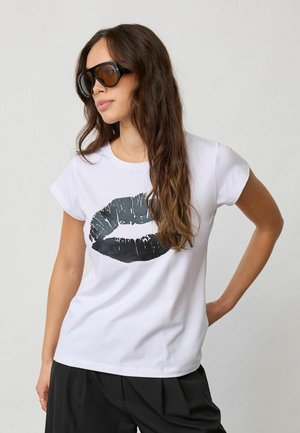 Donna con lunghi capelli castani che indossa occhiali da sole neri oversize e una T-shirt bianca con stampa labbra nere, abbinata a pantaloni neri, in posa davanti a uno sfondo grigio.