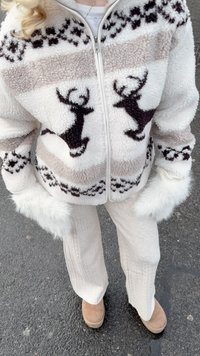 Witte fleecejas met rendiermotieven en zwarte accenten, voorzien van een ritssluiting aan de voorkant en witte nepvachtmanchetten. Gecombineerd met licht crèmekleurige, geribbelde broek en beige laarzen.