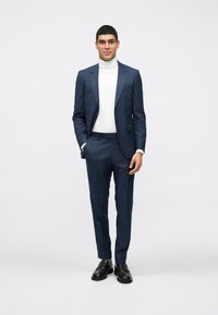Ναυτικό μπλε κοστούμι με στενή εφαρμογή, σε συνδυασμό με λευκό φανελάκι. Μαύρες δερμάτινες παπούτσια και διακριτική λεπτομέρεια τσέπης στο σακάκι.