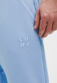 Světlé modré tepláky z měkké látky s vyšívaným logem. Design zahrnuje boční kapsy a volný střih.