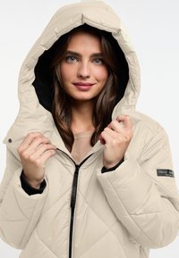Beige gewatteerde pufferjack met een grote capuchon, ritssluiting en zwarte accenten. Heeft een gestructureerde voering en een schouderpatch logo.