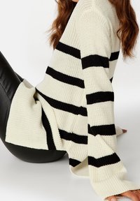 Brede gebreide trui in roomkleur met gedurfde zwarte horizontale strepen. Loose fit, lange mouwen met geribde manchetten en textiele stof.