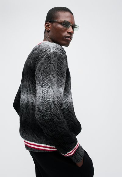 Paksu kudumiga halli gradient-sweater, millel on tekstuuriga nöörmuster. Omab ribikoes äärt, kus on punased ja valged triibud. Kantud prillidega.
