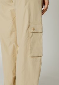 Pantaloni cargo beige con una texture liscia, dotati di una tasca laterale chiusa da un bottone. Design semplice con linee pulite e dettagli minimi.