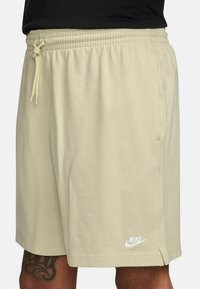 Vaaleanbeige urheilushortsit, joissa on joustava vyötärö ja kenguruvetoketju. Alhaalla vasemmalla puolella valkoinen Nike-logo. Silkkinen puuvillapinta.