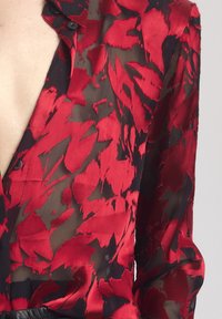 Rode en zwarte bloempatronen blouse van transparant materiaal, met een knoopsluiting bij de kraag en lange mouwen.