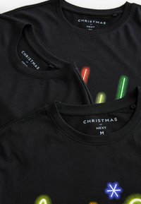 Dwa czarne bawełniane t-shirty nachodzą na siebie, mają kolorowe graficzne wzory w tematyce świątecznej. Metki informują "CHRISTMAS at NEXT" oraz rozmiar "M."