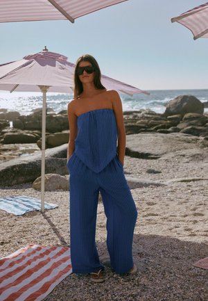 Kvinde iført blå strapless buksedragt og solbriller, der står på en klippefyldt strand under lyserøde stribede parasoller nær havet.