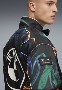 Svart racingjacka med färgglada mönstrade detaljer, BMW-logotyp på ryggen och en texturerad yta med kontrasterande vit piping.