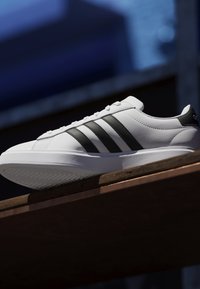 Weißer Sneaker mit schwarzen Streifen und strukturierter Sohle, auf einer Holzfläche vor einem dunkelblauen verschwommenen Hintergrund platziert.