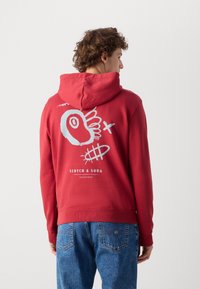 Red hooded sweatshirt with an oversized graphic design in light blue on the back. Features a front kangaroo pocket and ribbed cuffs.