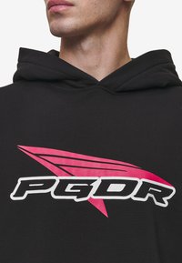 Felpa nera con cappuccio caratterizzata da un logo bianco e rosa ben visibile, accompagnato da un grafico stilizzato. Il materiale sembra morbido, con una vestibilità classica e cappuccio regolabile.