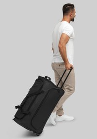 Sac de sport noir à roulettes avec des poignées robustes, tissu lisse et zippers. Comprend un grand compartiment principal et des roues robustes pour la mobilité.