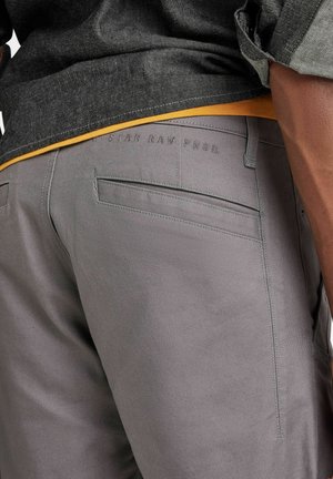 Gray pants with a smooth texture, featuring a back pocket and embossed branding, paired with a black shirt and contrasting yellow undershirt.