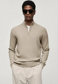 Beige ribbed sweater with a zip collar and long sleeves, paired with light trousers. The model wears dark sunglasses.