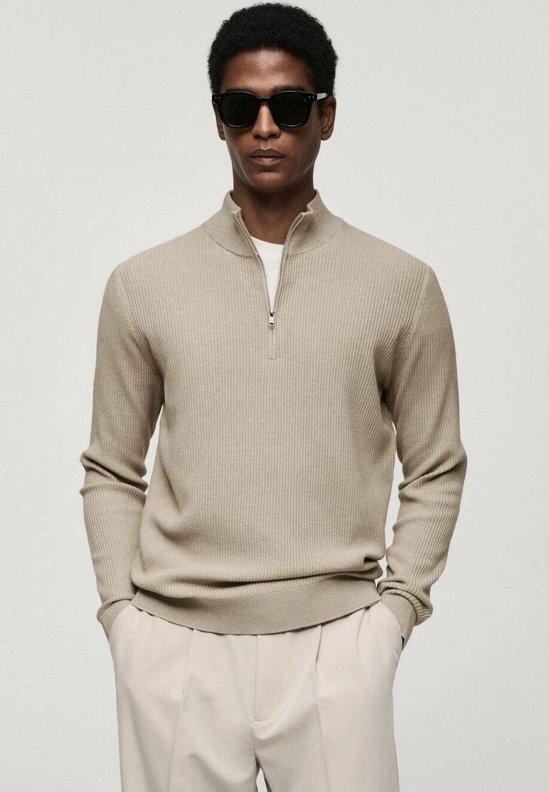 Beige ribbed sweater with a zip collar and long sleeves, paired with light trousers. The model wears dark sunglasses.