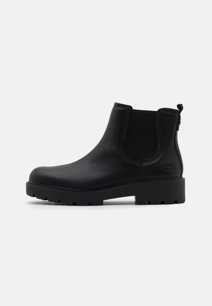 Schwarzer Chelsea-Lederstiefel mit elastischen Seitenpartien, grober Sohle, Fersenschlaufe und UGG-Logo an der Ferse, im Profil gezeigt.