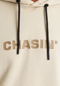 Beige kapucnis felső, texturált felülettel, nagy barna "CHASIN'" felirattal és fekete zsinórokkal, műanyag végződésekkel.