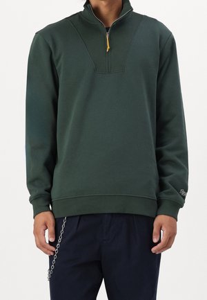 Mężczyzna ubrany w ciemnozielony sweter z zamkiem do ćwiartki i prążkowanym kołnierzem oraz mankietami, w połączeniu z ciemnymi spodniami i dodatkiem w postaci łańcucha.