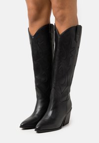 Zwarte leren cowboylaarzen met een spitse neus, hoge schacht en gevlochten details. Voorzien van een chunky hak en decoratieve steekaccenten.