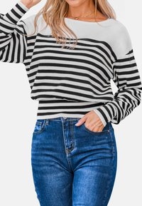 Mujer vestida con un suéter de manga larga de rayas blanco y negro y jeans azules, con una mano en el bolsillo.