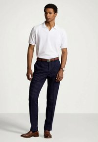 Mann iført hvit kortermet polo, marineblå stripebukser, brun belte, brune lærsko, og klokke, som står mot en ensfarget bakgrunn.