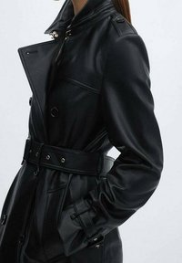 Trench-coat en cuir noir avec taille ceinturée, poignets boutonnés et large col, porté par une personne les mains dans les poches.