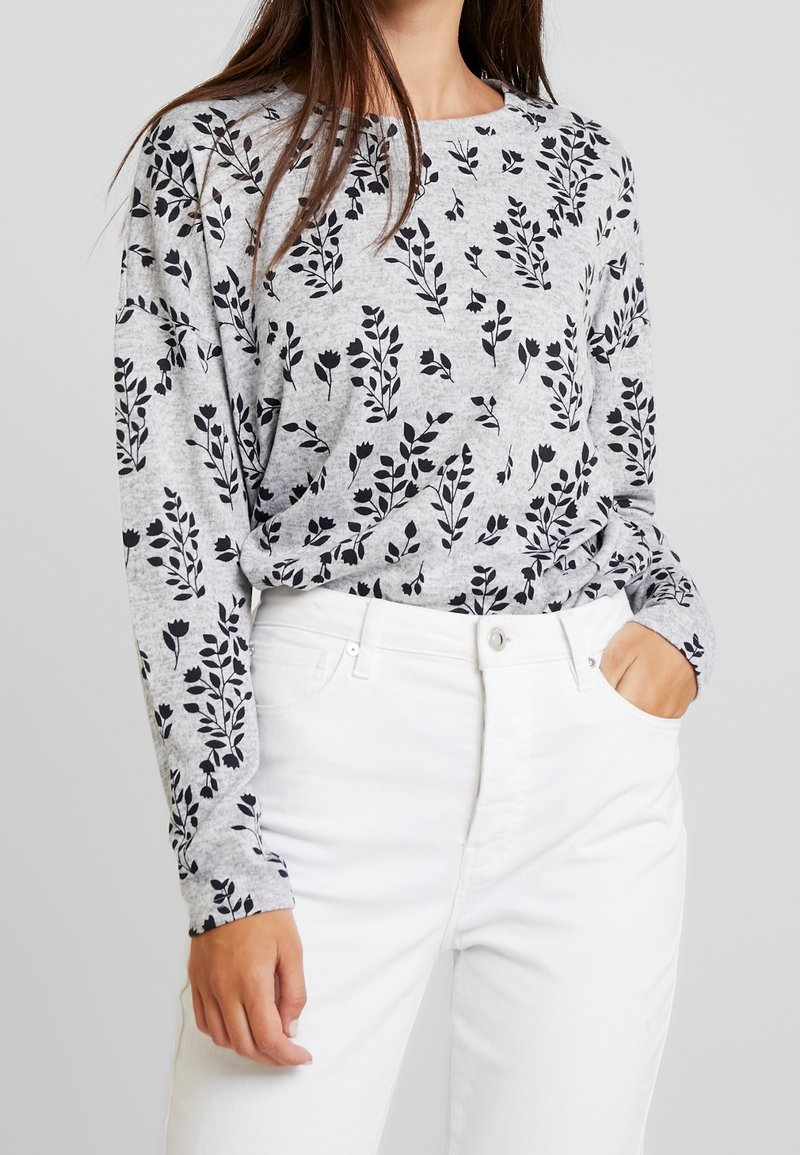 Femme portant un haut gris à manches longues avec un imprimé floral noir et un pantalon blanc taille haute, une main dans la poche, fond neutre.