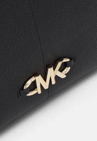 Sac à main en cuir noir avec une surface texturée, mettant en avant un accent en or avec le logo "CMK" et des anses en cuir noir.