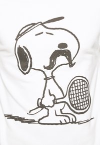 Camiseta blanca con un contorno negro de un perro de dibujos animados sosteniendo una raqueta de tenis, con un prominente diseño de bigote y gorra.