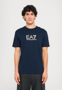 Tamsiai mėlyna medvilninė marškinėliai su apvaliu iškirpimu. Priekyje yra kontrastinės šviesios spalvos logotipas "EA7 EMPORIO ARMANI".