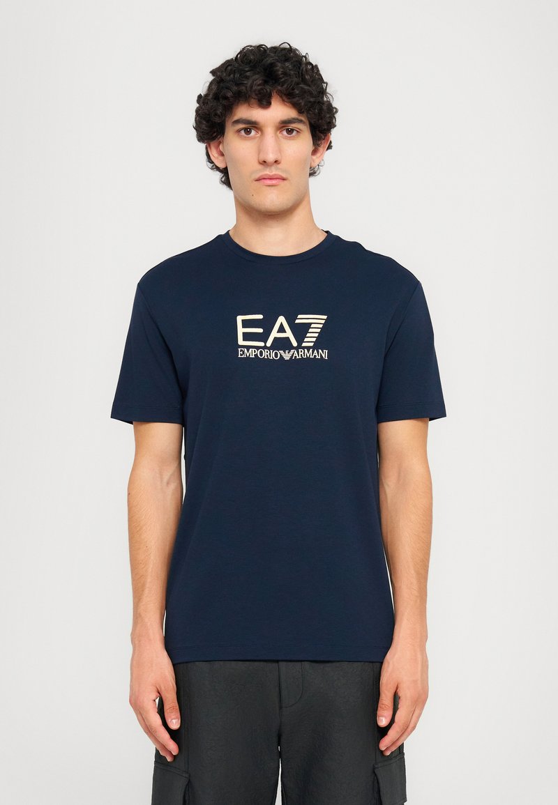 Tamsiai mėlyna medvilninė marškinėliai su apvaliu iškirpimu. Priekyje yra kontrastinės šviesios spalvos logotipas "EA7 EMPORIO ARMANI".
