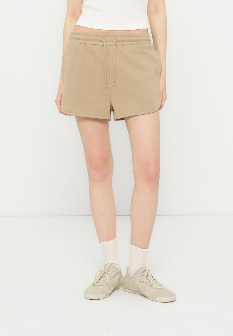 Femme portant un short beige à cordon, un top court blanc, des chaussettes blanches et des baskets beiges, debout devant un fond blanc uni.