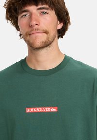 Zöld pamut póló, piros téglalap alakú logóval, amelyen a "QUIKSILVER" felirat és egy kis hegyikont látható. Klasszikus kerek nyakú fazon.